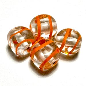 May include: Four clear glass beads with orange stripes. The beads are round and have a hole through the center for stringing. The orange stripes wrap around the beads, creating a decorative pattern. These beads are suitable for jewelry making.