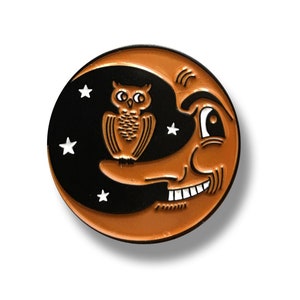 Pode incluir: Um broche de esmalte preto e laranja com uma lua crescente com um rosto sorridente e uma coruja empoleirada no topo. A lua tem estrelas brancas.
