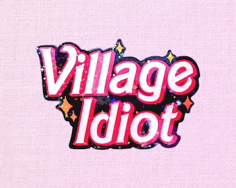 May include: A sticker with the words "Village Idiot" in white, outlined in red, and set against a black background. The sticker has a holographic, glittery effect and is decorated with yellow stars. The background is a light pink.