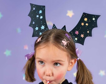 May include: A young girl wearing a black bat-wing headband with star cutouts and spider hair clips. She is drinking from a red and white striped straw with a bat-shaped decoration. The background is a light purple with star-shaped confetti.