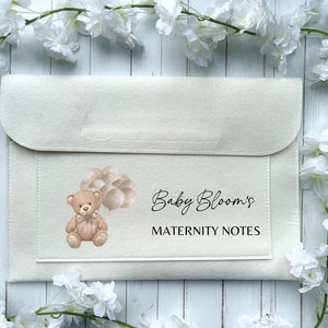 May include: A beige felt folder with a pocket on the front. The folder has a brown teddy bear with balloons and the text "Baby Bloom's Maternity Notes" printed on the front.
