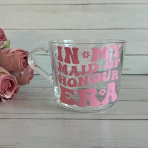 May include: A clear glass mug with a handle, featuring the pink text "IN MY MAID OF HONOUR ERA" in a retro font. The mug is decorated with flower accents. Pink roses are in the background.