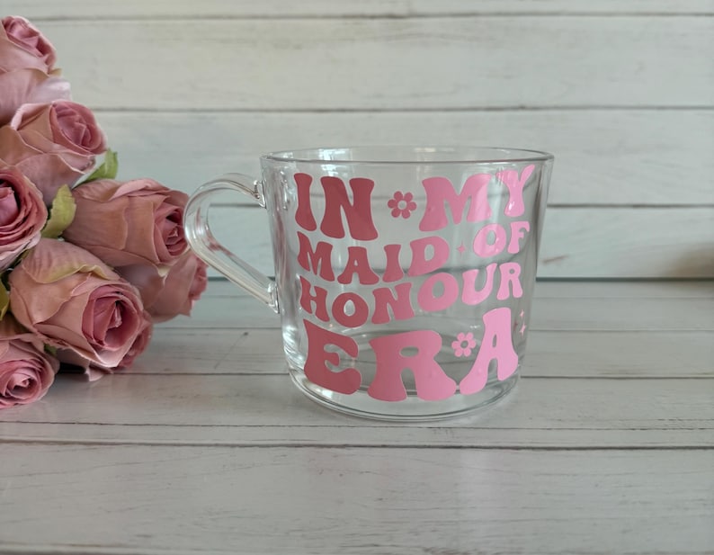 May include: A clear glass mug with a handle, featuring the pink text "IN MY MAID OF HONOUR ERA" in a retro font. The mug is decorated with flower accents. Pink roses are in the background.
