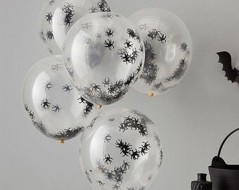 May include: Five clear balloons filled with black spider confetti. The balloons are clustered together, with some partially overlapping. A black bat decoration and a cauldron are visible in the background, suggesting a Halloween theme.