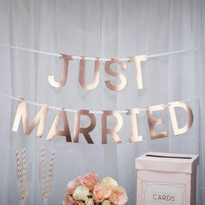 Può includere: Uno striscione "JUST MARRIED" in oro rosa è appeso su uno sfondo bianco. Sotto, un bouquet di rose rosa e crema, calici di champagne e cannucce a righe. È visibile anche una scatola per biglietti, suggerendo un matrimonio.