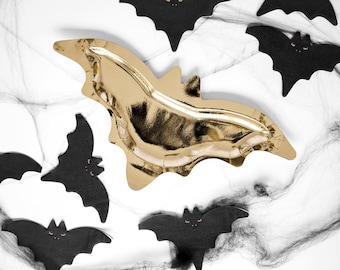 May include: A gold foil bat-shaped serving tray is the focal point, surrounded by black bat cutouts and a spiderweb design. The Halloween-themed decorations are set against a white background, creating a festive and spooky atmosphere.