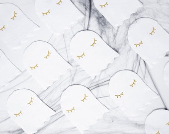May include: White ghost-shaped paper napkins with gold eyelashes, arranged on a gray and white surface with black spiderweb accents. These napkins are perfect for a Halloween or spooky-themed party.