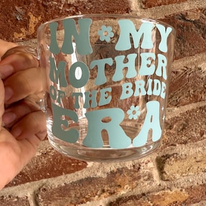 May include: Clear glass mug with a handle, featuring the text "IN MY MOTHER OF THE BRIDE ERA" in light blue, retro-style lettering. A floral graphic accents the text. The mug is held against a brick wall.