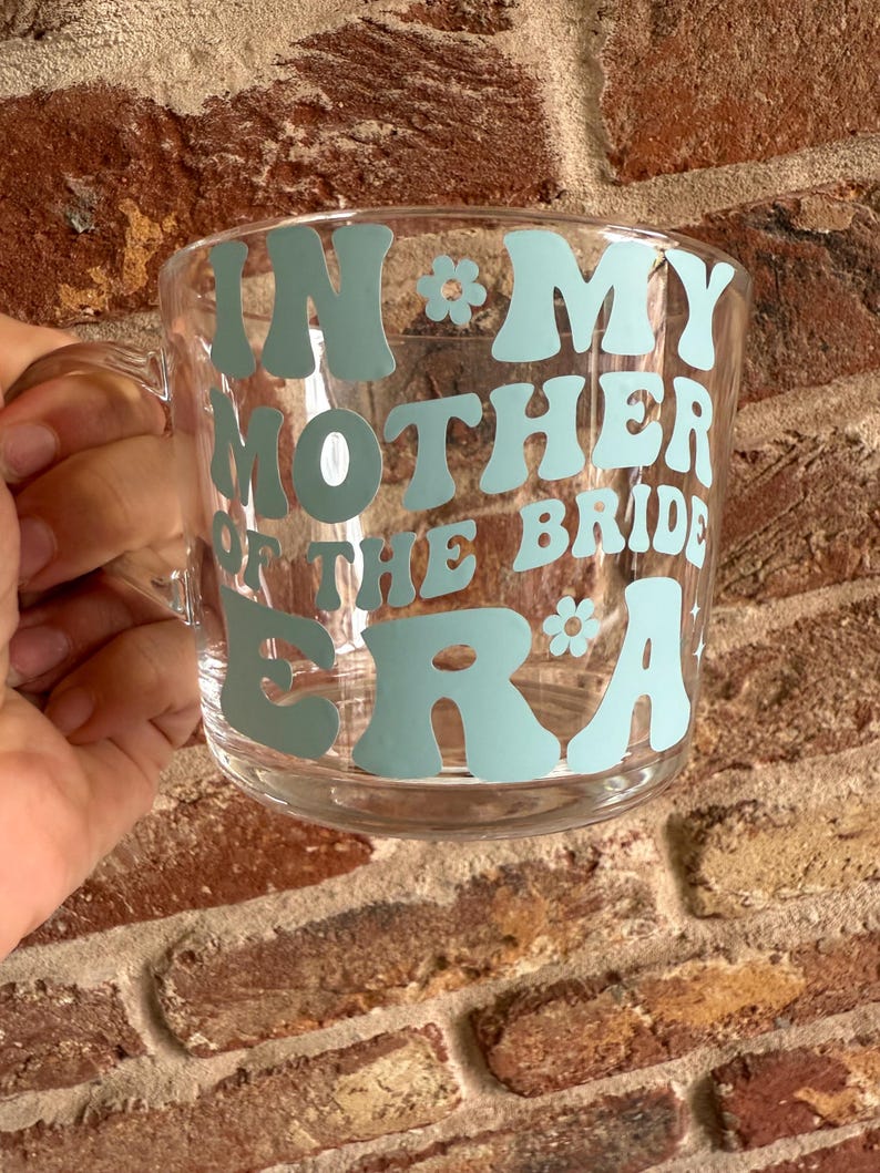 May include: Clear glass mug with a handle, featuring the text "IN MY MOTHER OF THE BRIDE ERA" in light blue, retro-style lettering. A floral graphic accents the text. The mug is held against a brick wall.