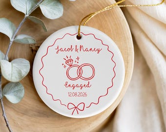 May include: A round white ceramic ornament featuring a red scalloped border and a gold hanging string. The design includes the text Jacob & Nancy, Engaged, and 12.08.2026, accompanied by a red line drawing of two interlocking rings and a diamond.