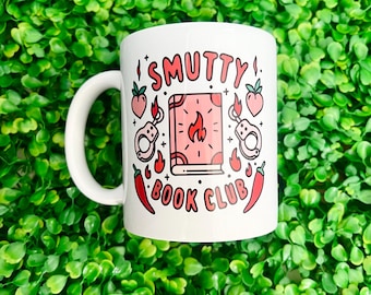 Könnte beinhalten: Ein weißer Keramikbecher mit einem grafischen Design und dem Text SMUTTY BOOK CLUB in roten Buchstaben. Die Illustration zeigt ein rosa Buch mit einem Flammensymbol, Handschellen, zwei Pfirsichen und zwei roten Chilischoten, umgeben von kleinen schwarzen Sternen.