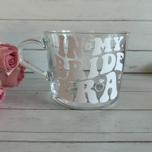 May include: Clear glass mug with a handle, featuring the text "IN MY BRIDE ERA" in white and light pink lettering. The design includes a ring and flower accents. Pink roses are visible in the background.