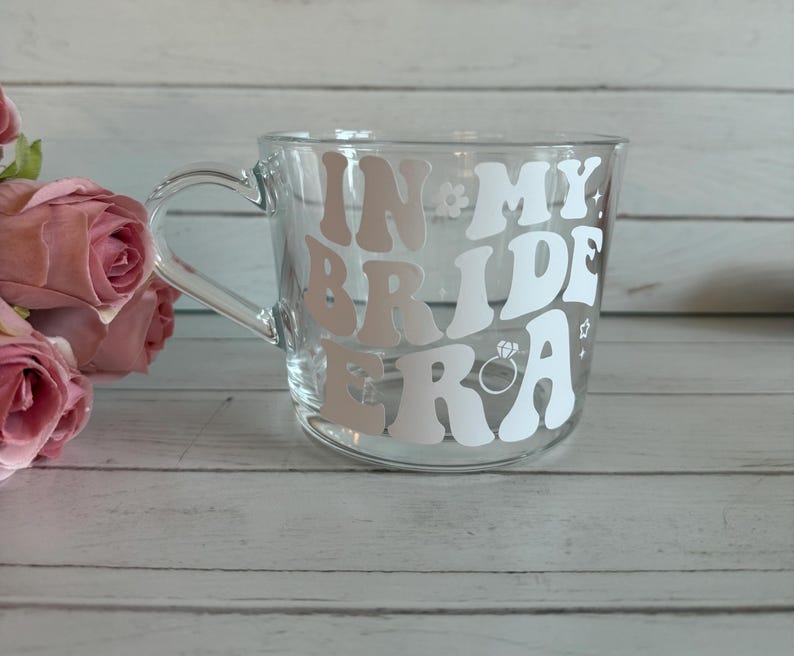 May include: Clear glass mug with a handle, featuring the text "IN MY BRIDE ERA" in white and light pink lettering. The design includes a ring and flower accents. Pink roses are visible in the background.