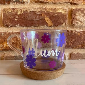 May include: A clear glass mug with a cork coaster. The mug has a white handle and is decorated with purple and lavender flowers. The word "Mum" is written in white on the mug.