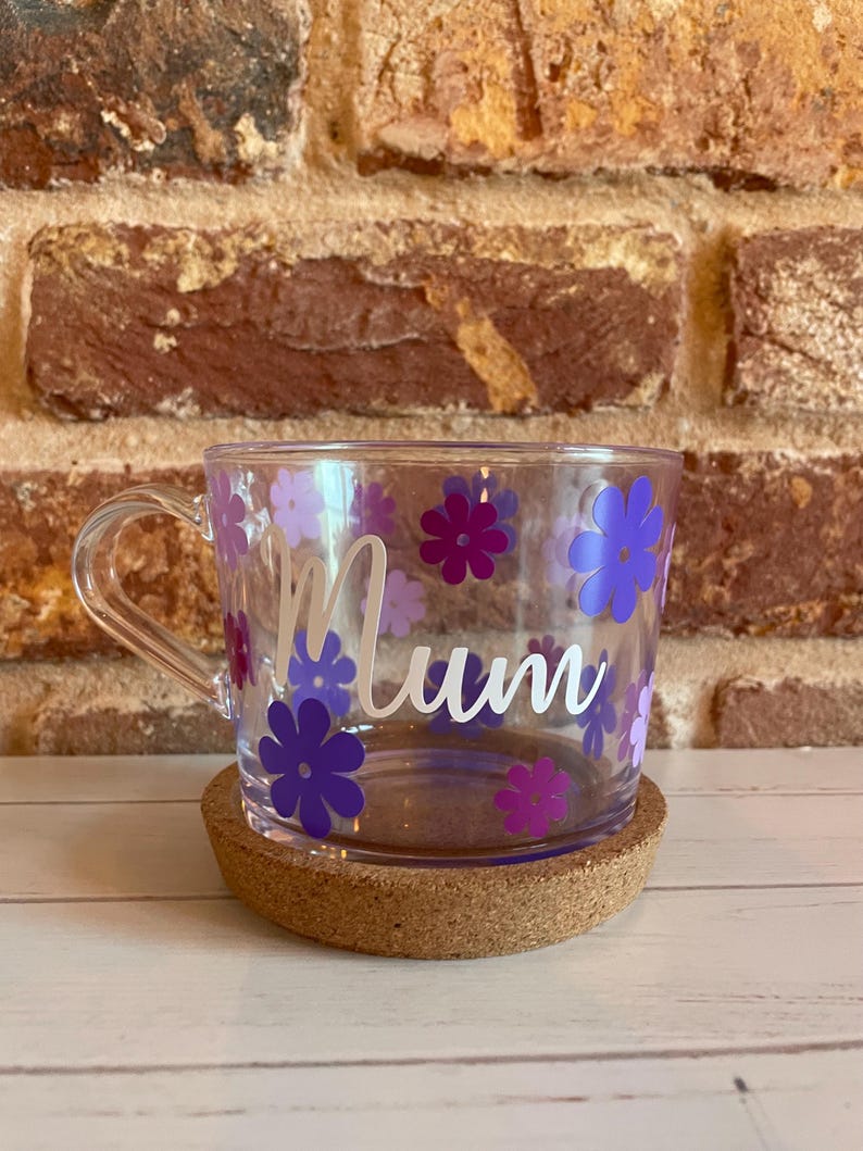 May include: A clear glass mug with a cork coaster. The mug has a white handle and is decorated with purple and lavender flowers. The word "Mum" is written in white on the mug.