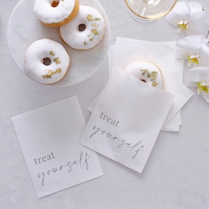 May include: An overhead shot shows white iced doughnuts with gold leaf, a marble cake stand, and white paper bags printed with "treat yourself". Napkins and white orchids are also visible, creating an elegant display.