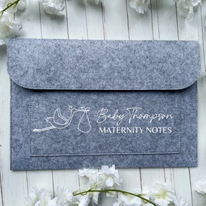 May include: A gray felt maternity notes folder with a flap closure. The folder features a white stork carrying a baby and the text "Baby Thompson MATERNITY NOTES". The folder is surrounded by white flowers.