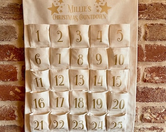 May include: A beige fabric Christmas advent calendar with 25 numbered pockets. The calendar has the text "Millie's Christmas Countdown" in gold script, with star accents. It hangs from a wooden dowel and a natural rope.