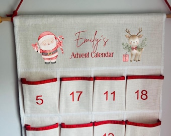 May include: A beige fabric advent calendar with red trim and pockets numbered 1-24. The top features a watercolor Santa Claus and reindeer illustration, with the text "Emily's Advent Calendar" in red script. The calendar hangs from a wooden dowel with red rope.