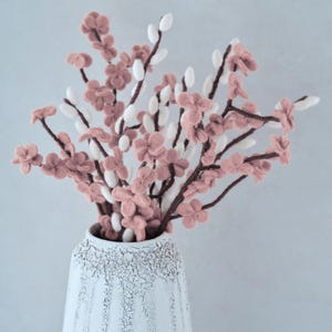 May include: A white ceramic vase with a textured surface holds a bouquet of pink and white felt flowers. The flowers are arranged in a loose, natural style.