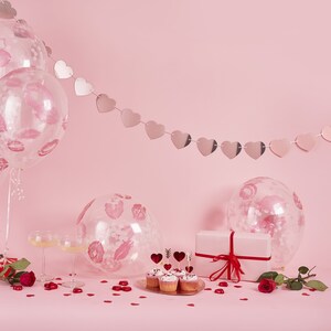 May include: A romantic scene with pink balloons printed with lip imprints, a heart-shaped garland, cupcakes, and a gift box tied with a red ribbon. Scattered red heart confetti and roses complete the Valentine's Day theme.