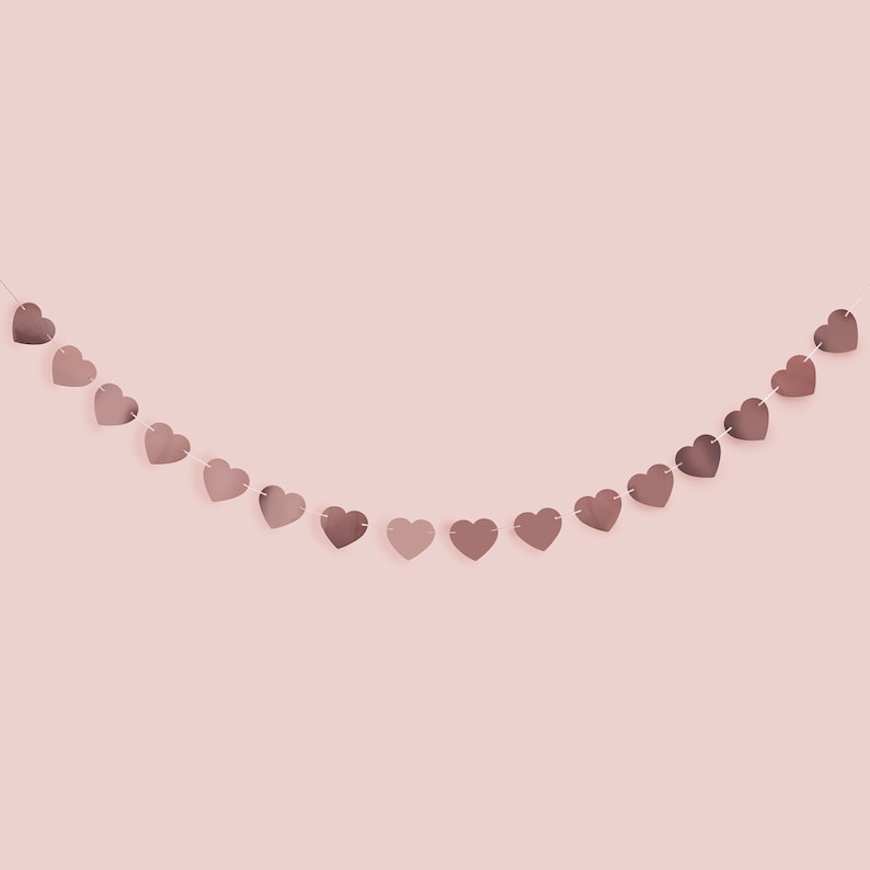 May include: A garland of rose gold heart-shaped cutouts strung together with white thread. The hearts are evenly spaced and hang against a soft pink background. The garland is slightly curved, creating a gentle arc.