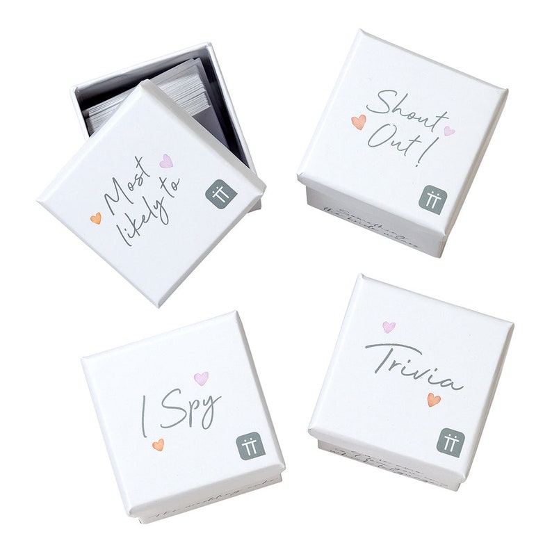 May include: Four white boxes with different game titles printed on them. The boxes are labeled "Most Likely To", "Shout Out!", "I Spy", and "Trivia". Each box has a small pink heart and a small orange heart printed on it.