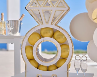 May include: A white and gold diamond ring-shaped decoration filled with gold-colored spheres. The ring sits on a white table with champagne flutes and balloons in the background. A festive decoration for a wedding or engagement.