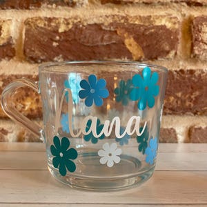 May include: A clear glass mug with a white handle and blue and white flower decals. The mug has the word "Nana" written in white cursive lettering.
