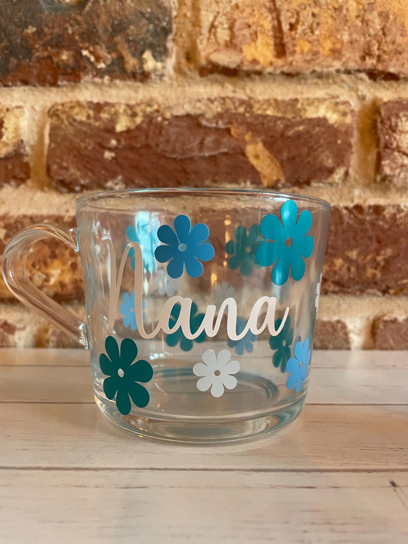 May include: A clear glass mug with a white handle and blue and white flower decals. The mug has the word "Nana" written in white cursive lettering.