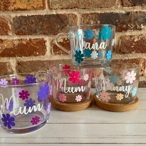 May include: Four clear glass mugs with floral designs and personalized names. The mugs are decorated with colorful flowers and the names "Nana", "Mum", "Mum", and "Nanny" are written in white lettering.