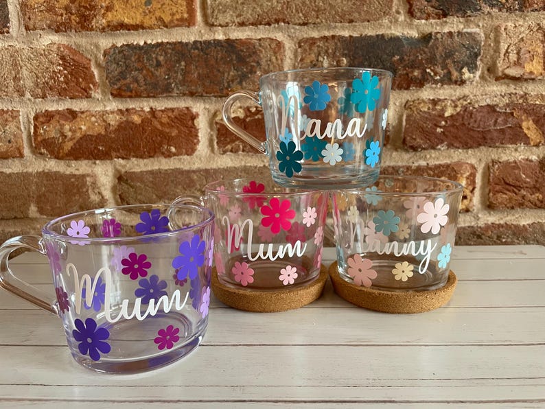 May include: Four clear glass mugs with floral designs and personalized names. The mugs are decorated with colorful flowers and the names "Nana", "Mum", "Mum", and "Nanny" are written in white lettering.