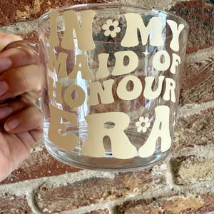 May include: Clear glass mug with a handle, featuring the text "IN MY MAID OF HONOUR ERA" in a retro font. The text is in a light beige color, with small flower graphics. The mug is held by a person.
