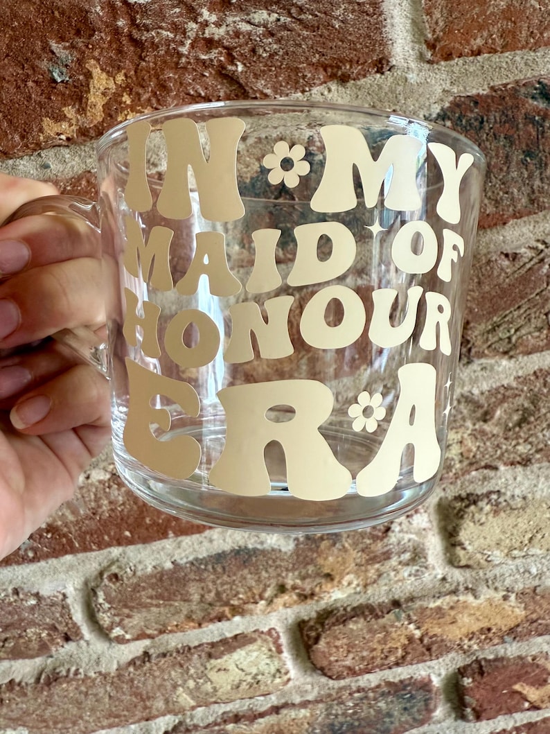 May include: Clear glass mug with a handle, featuring the text "IN MY MAID OF HONOUR ERA" in a retro font. The text is in a light beige color, with small flower graphics. The mug is held by a person.