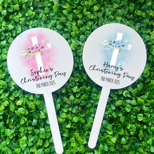 May include: Two white round signs with a white cross and floral design. The sign on the left is pink with purple flowers and says "Sophie's Christening Day 2nd March 2025". The sign on the right is blue with blue flowers and says "Harry's Christening Day 2nd March 2025".
