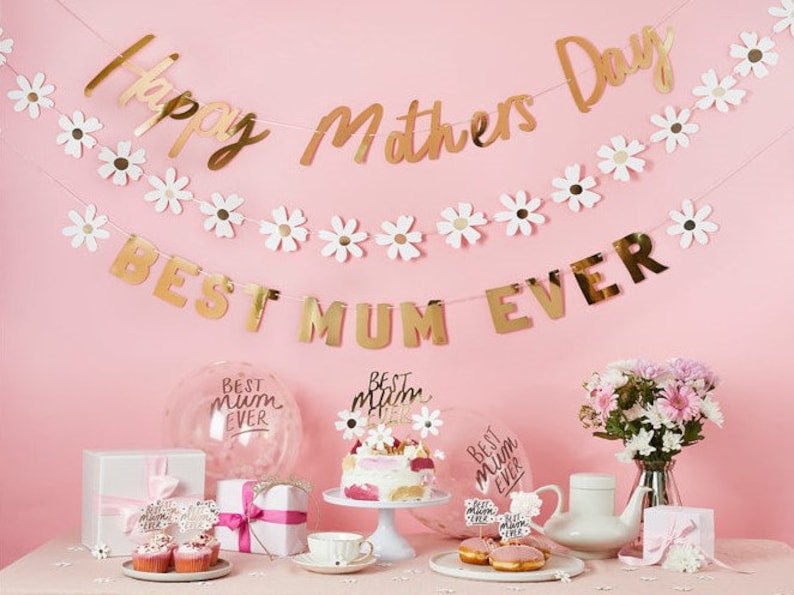 May include: A festive Mother's Day display with gold banners reading "Happy Mothers Day" and "Best Mum Ever." The scene includes a cake, cupcakes, gifts, balloons, and flowers, all set against a pink backdrop.