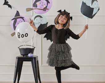 May include: A young person in a black bat costume with a layered tulle skirt holds Halloween-themed balloons. The balloons feature witch and vampire illustrations, and the word "Boo". A black cauldron and a purple pumpkin sit on a black stool.