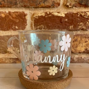 May include: A clear glass mug with a cork coaster. The mug has a white floral design with the word "Nanny" written on it.