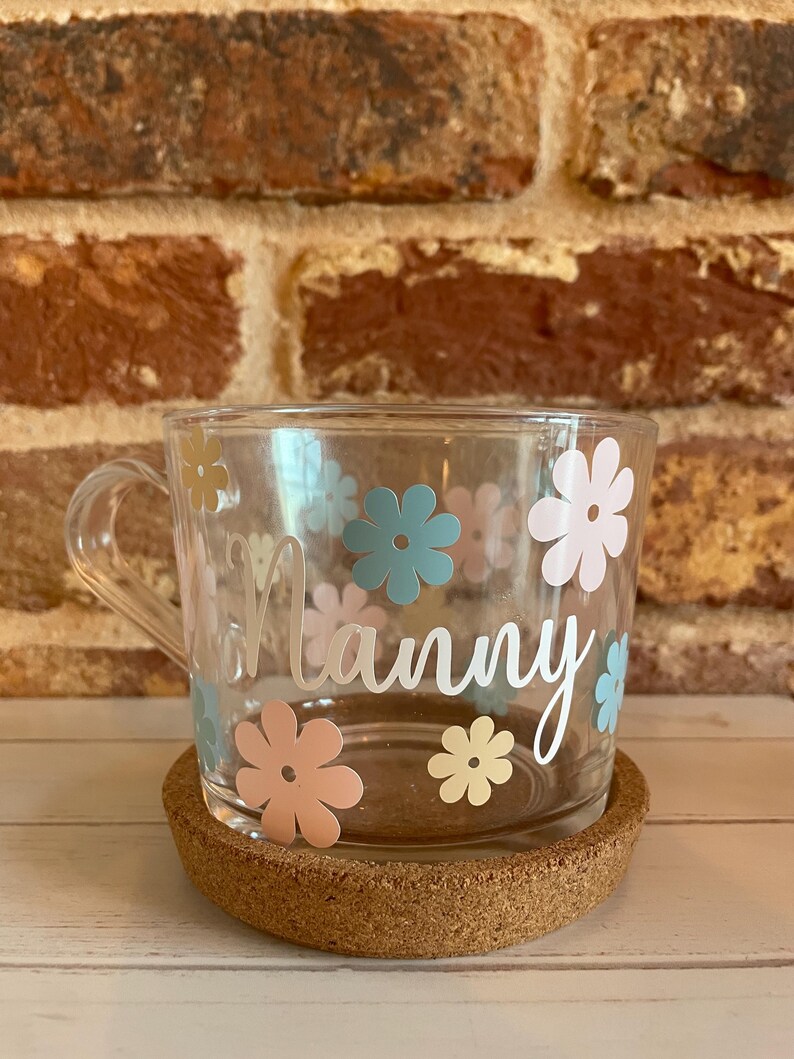 May include: A clear glass mug with a cork coaster. The mug has a white floral design with the word "Nanny" written on it.