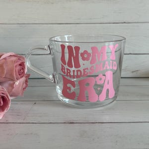 May include: A clear glass mug with a handle, featuring the text "IN MY BRIDESMAID ERA" in pink lettering. The mug is set against a light wood background, with pink roses in the foreground.