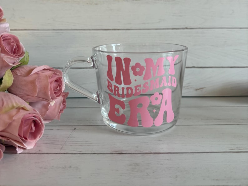 May include: A clear glass mug with a handle, featuring the text "IN MY BRIDESMAID ERA" in pink lettering. The mug is set against a light wood background, with pink roses in the foreground.
