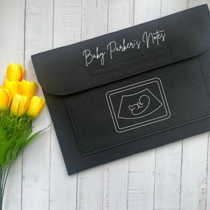 May include: A black felt document holder with the text "Baby Parker's Notes" in a script font. The front pocket features a white outline of a sonogram image. A bouquet of yellow tulips sits to the left of the document holder.
