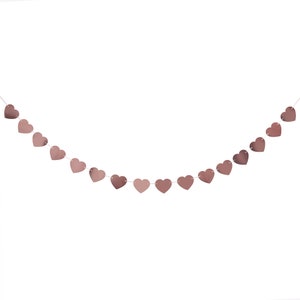 May include: A string of rose gold heart-shaped decorations hangs against a white background. The hearts are connected by a thin, white string, creating a festive garland. The hearts have a metallic sheen, reflecting light.