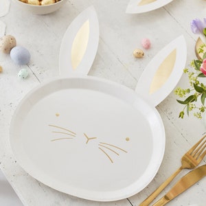 May include: White paper plates in the shape of a bunny face with gold detailing. The plates feature bunny ears and gold whiskers. Easter-themed plates are on a white wooden surface with Easter eggs and gold cutlery.