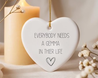 May include: A white heart-shaped ceramic ornament hanging from a gold string. The text reads: EVERYBODY NEEDS A GEMMA IN THIER LIFE, followed by a small hand-drawn heart outline. The ornament has a smooth, glossy finish.