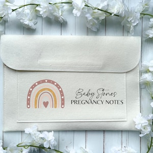 May include: A cream-coloured felt pregnancy notes holder with a flap closure. The front displays a rainbow design in pink, peach, and yellow, and the text "Baby Stones PREGNANCY NOTES".