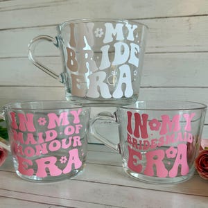May include: Three clear glass mugs with handles, each featuring text in a retro font. The top mug says "In My Bride Era" in white and tan. The bottom mugs say "In My Maid of Honour Era" and "In My Bridesmaid Era" in pink.