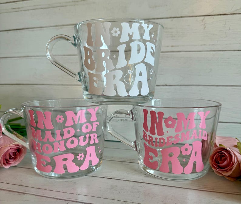 May include: Three clear glass mugs with handles, each featuring text in a retro font. The top mug says "In My Bride Era" in white and tan. The bottom mugs say "In My Maid of Honour Era" and "In My Bridesmaid Era" in pink.