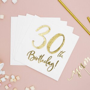 Puede incluir: Servilletas de papel blancas con texto dorado "30th Birthday!". Pajitas doradas, un adorno para pastel "yum" y una taza rosa a rayas con malvaviscos sobre un fondo rosa.