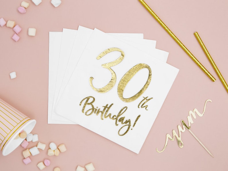 Puede incluir: Servilletas de papel blancas con texto dorado "30th Birthday!". Pajitas doradas, un adorno para pastel "yum" y una taza rosa a rayas con malvaviscos sobre un fondo rosa.